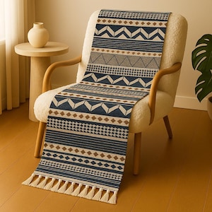 May include: A navy blue and tan patterned table runner with geometric designs, including triangles, diamonds, and stripes. The runner has a cream-colored fringe at the bottom and is draped over a white armchair.