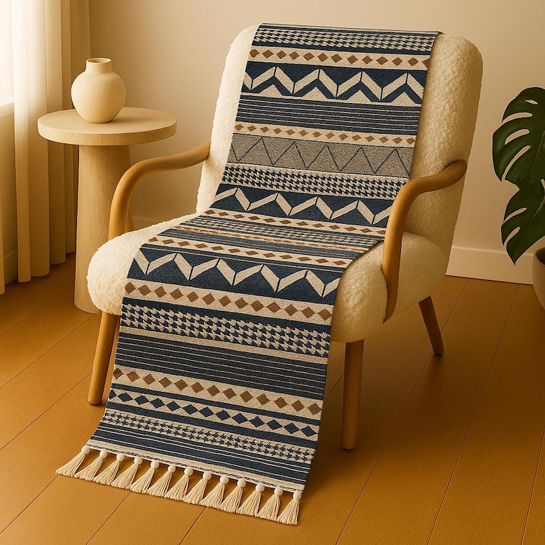 May include: A navy blue and tan patterned table runner with geometric designs, including triangles, diamonds, and stripes. The runner has a cream-colored fringe at the bottom and is draped over a white armchair.