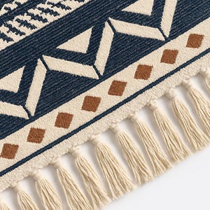 May include: Close-up of a woven rug with a geometric pattern. The rug features a navy blue background with cream and brown geometric shapes. The edge is trimmed with cream-colored tassels, adding a decorative element to the rug.