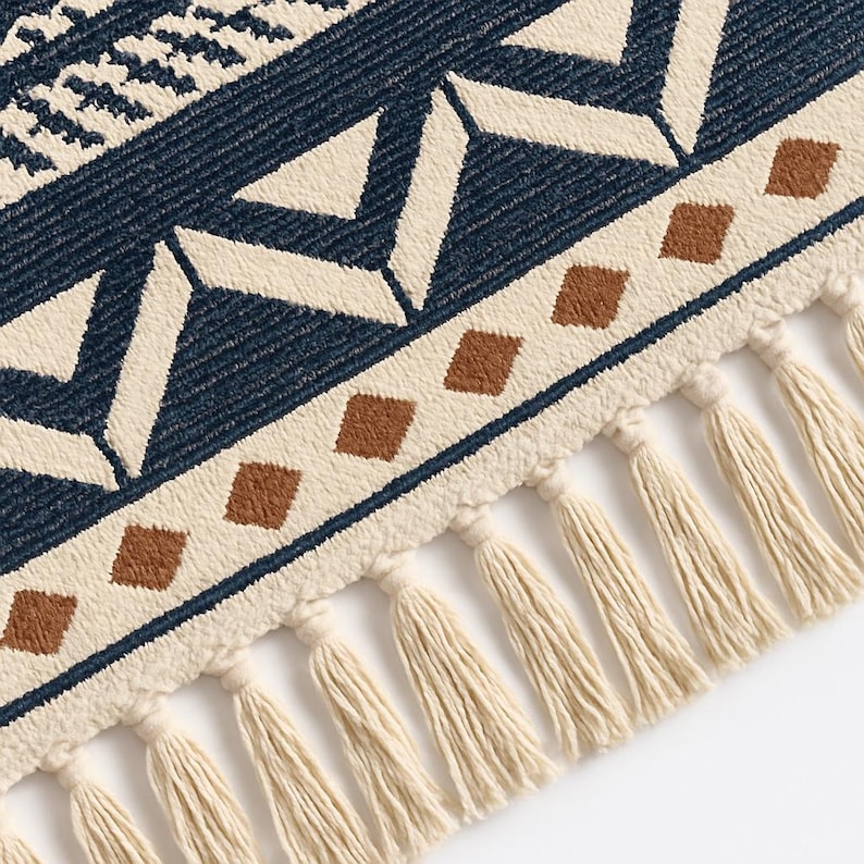 May include: Close-up of a woven rug with a geometric pattern. The rug features a navy blue background with cream and brown geometric shapes. The edge is trimmed with cream-colored tassels, adding a decorative element to the rug.