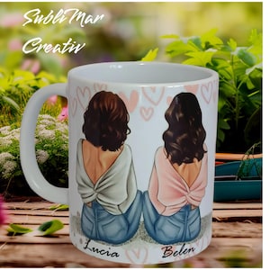 May include: White ceramic mug featuring a design of two women with brown hair wearing sweaters and jeans. The mug has pink hearts and the names "Lucia" and "Belen" written on the bottom.