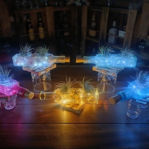 May include: Several repurposed glass bottles, each with a cork, are transformed into planters. Each bottle holds a small plant and is illuminated with colorful LED lights. The bottles rest on small wooden platforms supported by glasses.