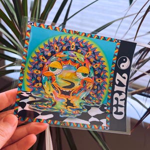 Può includere: Una colorata stampa artistica con un design psichedelico raffigurante un volto sorridente e molteplici occhi. L'opera d'arte ha uno sfondo blu con un bordo arcobaleno e un motivo a scacchiera in bianco e nero nella parte inferiore. La parola "GRIZ" è stampata verticalmente su un'etichetta nera.