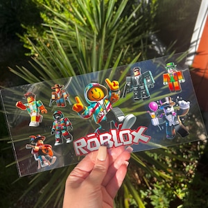 May include: A clear sticker sheet featuring various Roblox character designs. The sheet includes characters in different outfits, a smiley face character, and the Roblox logo in red and white.