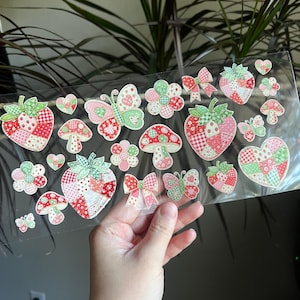 May include: A sheet of decorative stickers featuring strawberries, butterflies, flowers, mushrooms, hearts, and bows. The stickers have a patchwork design in shades of red, pink, and green. The stickers are held in a person's hand.