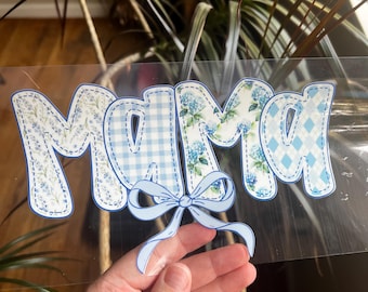 May include: A clear sticker with the word "MAMA" in a whimsical font. The letters are decorated with blue floral, gingham, and diamond patterns. A light blue bow is centered below the word.