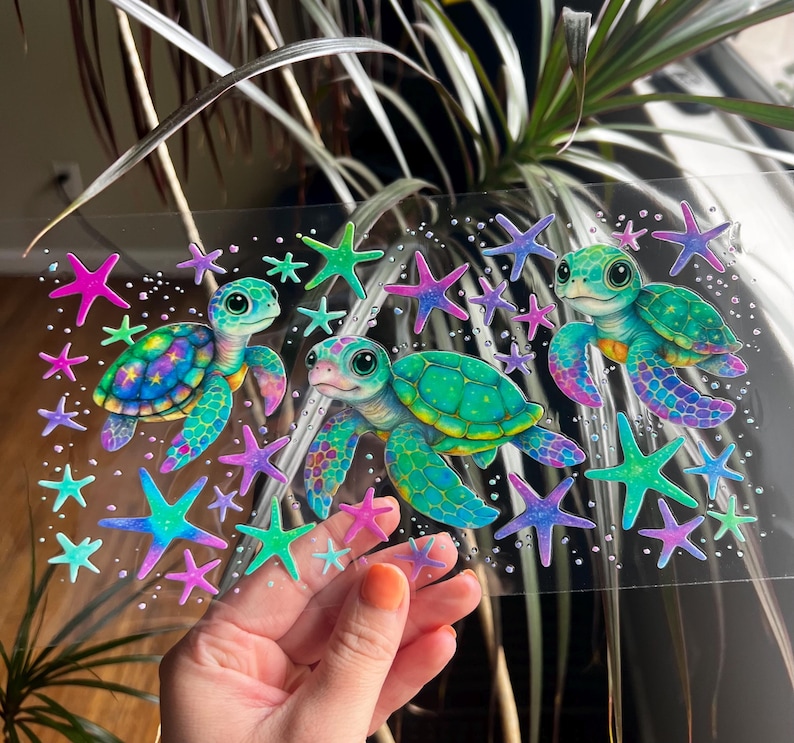 Pu&ograve; includere: Un adesivo trasparente con tre tartarughe marine dei cartoni animati in tonalit&agrave; di verde, blu e viola. Le tartarughe sono circondate da stelle marine colorate in rosa, verde e viola. L'adesivo &egrave; tenuto contro uno sfondo di piante verdi.