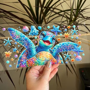 Pode incluir: Ilustração colorida de uma tartaruga marinha sorridente, rodeada por estrelas do mar, conchas e bolhas. A carapaça da tartaruga é uma mistura vibrante de azuis, verdes e laranjas. A imagem é segurada por uma mão.