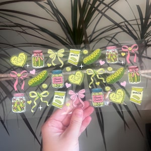 May include: A clear sticker sheet with pickle-themed illustrations. Designs include jars of pickles, pickle slices, heart-shaped pickles, bows, and snack bags. The color palette consists of green, yellow, pink, and blue.