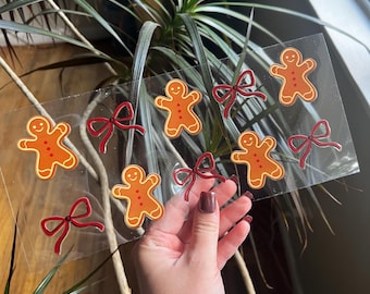 Op de afbeelding: Een doorzichtig vel met peperkoekmannetjesstickers en rode strikstickers. De peperkoekmannetjes zijn oranje met witte glazuurdetails. De strikken zijn dieprood van kleur. De stickers worden in de hand van een persoon gehouden.
