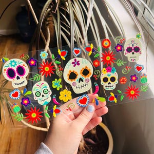 Puede incluir: Una hoja de pegatinas coloridas con tem&aacute;tica del D&iacute;a de Muertos con calaveras de az&uacute;car con dise&ntilde;os florales. Las pegatinas est&aacute;n sobre un fondo transparente y son perfectas para agregar un toque festivo a las manualidades, los regalos y la decoraci&oacute;n del hogar.