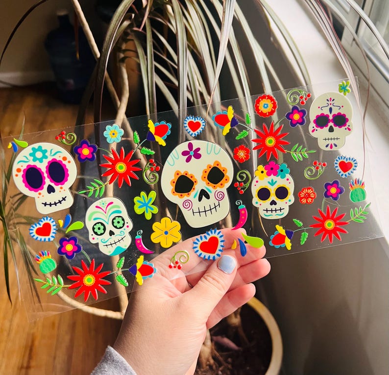 Puede incluir: Una hoja de pegatinas coloridas con tem&aacute;tica del D&iacute;a de Muertos con calaveras de az&uacute;car con dise&ntilde;os florales. Las pegatinas est&aacute;n sobre un fondo transparente y son perfectas para agregar un toque festivo a las manualidades, los regalos y la decoraci&oacute;n del hogar.