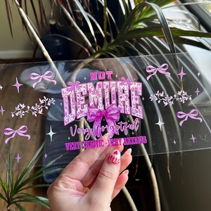 Puede incluir: Adhesivo transparente con el texto "NOT DEMURE" en una fuente decorativa, con las palabras "Very forgetful, Very chaotic, Very anxious" debajo. La pegatina tiene lazos rosas, estrellas y detalles florales.