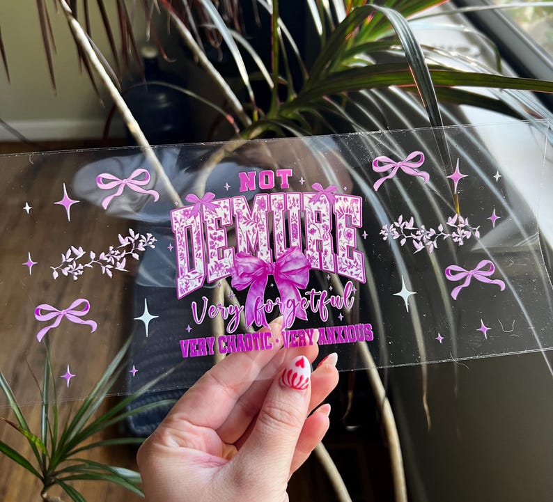 Puede incluir: Adhesivo transparente con el texto "NOT DEMURE" en una fuente decorativa, con las palabras "Very forgetful, Very chaotic, Very anxious" debajo. La pegatina tiene lazos rosas, estrellas y detalles florales.