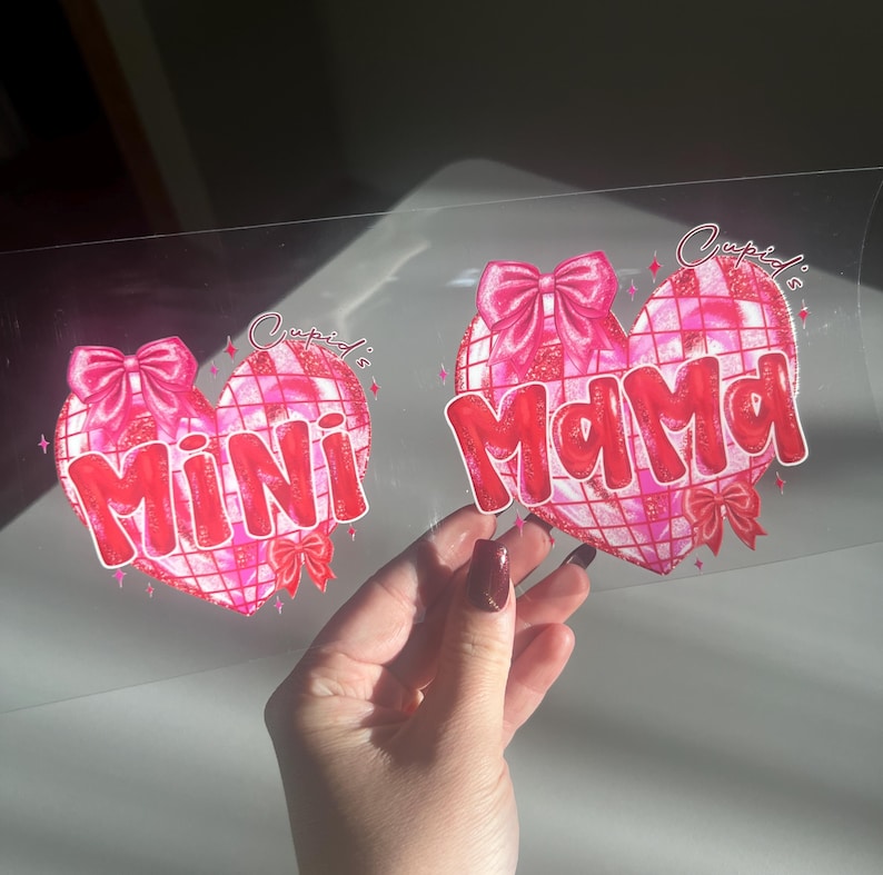 May include: Two pink heart-shaped designs with the words "Mini" and "Mama" in red, each adorned with a bow and a grid pattern. The text "Cupid's" is written above the hearts.