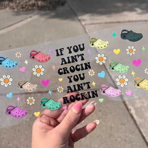 May include: A transparent sticker sheet with cartoon Crocs, daisies, hearts, and stars. The text says "If you ain't Crocin you ain't Rockin". The Crocs are in blue, pink, green, and yellow. A fun, colourful design.