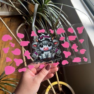 以下が含まれることがあります： 「HOT MESS」と書かれた帽子をかぶった漫画の牛が描かれた透明なステッカーシート。牛は白黒でピンクのアクセントが付いています。牛の周りにはピンクの牛柄の形があります。