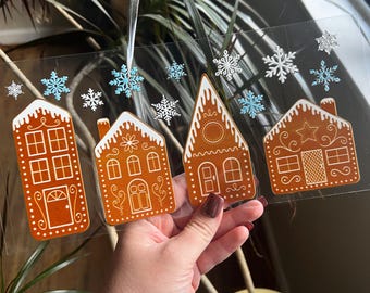 Op de afbeelding: Transparante kerstversiering met vier peperkoekhuisjes, witte glazuur en decoratieve details. De versiering heeft blauwe en witte sneeuwvlokaccenten en een transparant lint. Een feestelijke vakantiedecoratie.