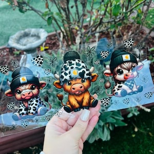 Puede incluir: Tres vacas de las Tierras Altas de dibujos animados con gorros Carhartt y trajes estampados. Las vacas están sentadas, una con un traje blanco y negro, otra con un gorro blanco y negro y la otra con una bufanda azul y blanca.