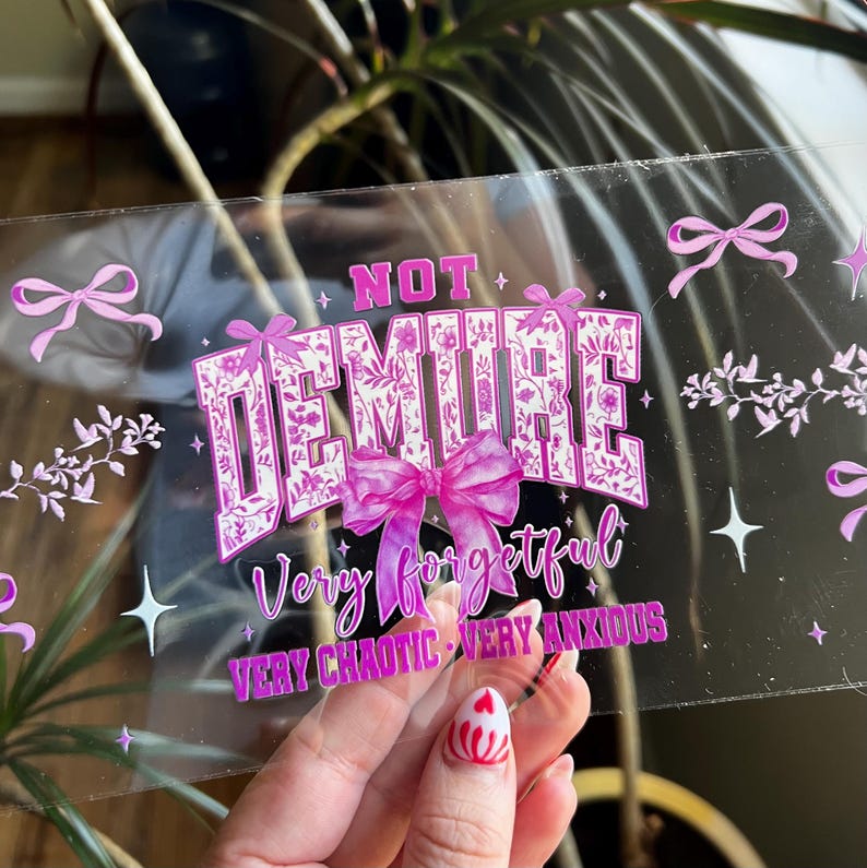 Puede incluir: Una pegatina transparente con las palabras "NOT DEMURE" en una fuente floral, con un gran lazo rosa. Debajo, el texto dice "Very forgetful, very chaotic, very anxious". Lazos y estrellas rosas decoran los bordes.