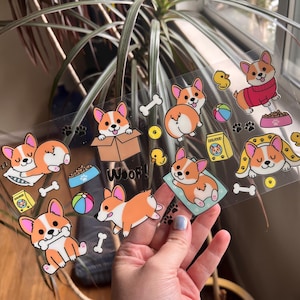 May include: A sheet of clear stickers featuring cartoon corgis in various poses, including sleeping, playing fetch, and wearing a red sweater. The stickers also include a bone, a ball, a bowl of dog food, and the word "Woof!" in black letters.