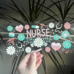 May include: A clear sticker with the word "NURSE" in pink letters, surrounded by floral and medical-themed illustrations. The design includes daisies, hearts, stethoscopes, and a syringe. The sticker is held in a person's hand.