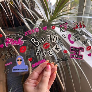 May include: A clear sheet adorned with stickers, including phrases like "Burn Book" and "That's so fetch." The stickers feature hearts, lips, and cartoon figures. A hand holds a small red sticker. The overall theme is a pop culture reference.