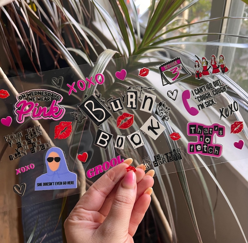 May include: A clear sheet adorned with stickers, including phrases like "Burn Book" and "That's so fetch." The stickers feature hearts, lips, and cartoon figures. A hand holds a small red sticker. The overall theme is a pop culture reference.