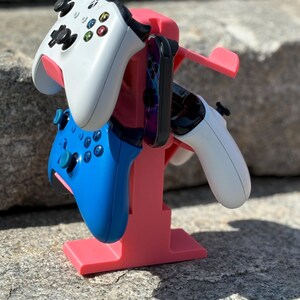 Puede incluir: Un soporte de pl&aacute;stico rosa sostiene tres mandos de videojuegos. Un mando es blanco, otro es azul y otro es blanco y negro. El soporte tiene un peque&ntilde;o estante para un tel&eacute;fono u otro dispositivo.