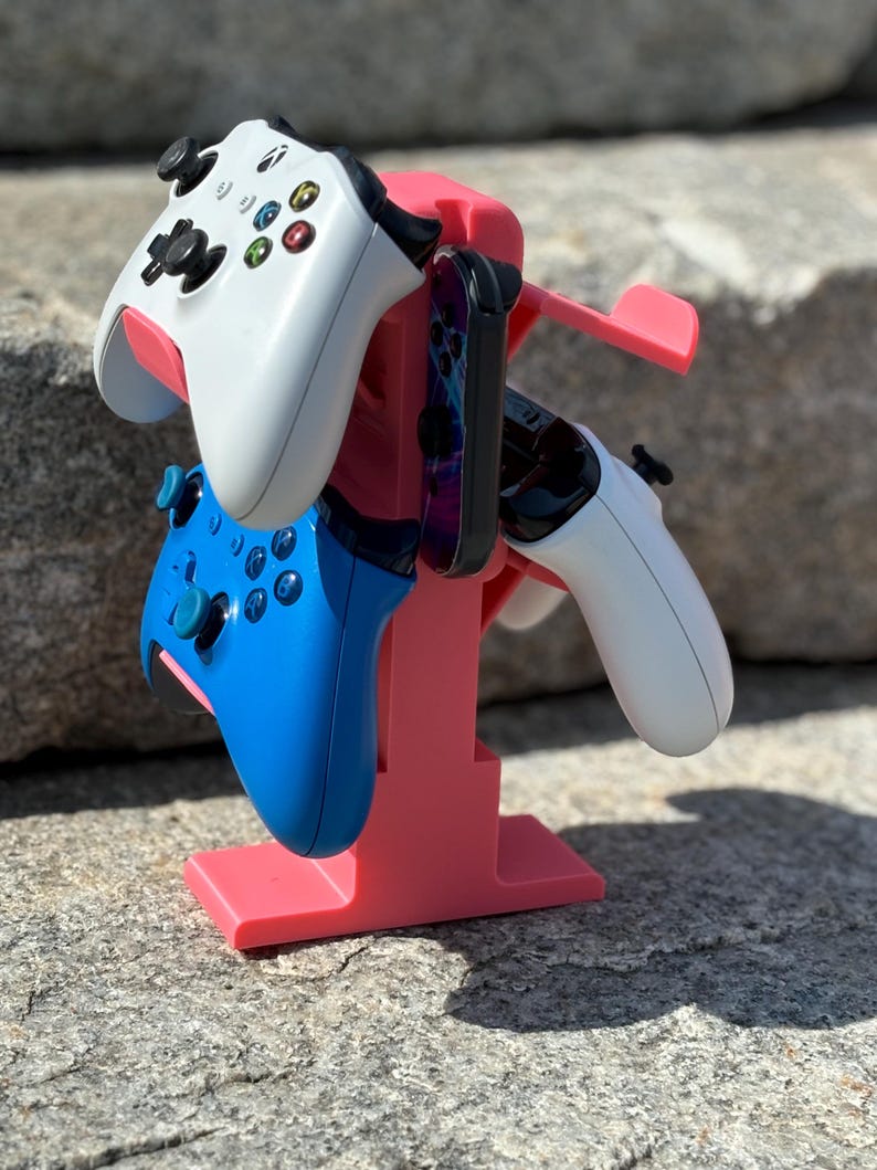 Puede incluir: Un soporte de pl&aacute;stico rosa sostiene tres mandos de videojuegos. Un mando es blanco, otro es azul y otro es blanco y negro. El soporte tiene un peque&ntilde;o estante para un tel&eacute;fono u otro dispositivo.