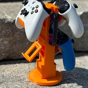 Puede incluir: Soporte de pl&aacute;stico naranja con tres mandos de videojuegos. El soporte tiene una base redonda y una estructura vertical con ranuras para sujetar los mandos. Los mandos son blancos, negros y azules.