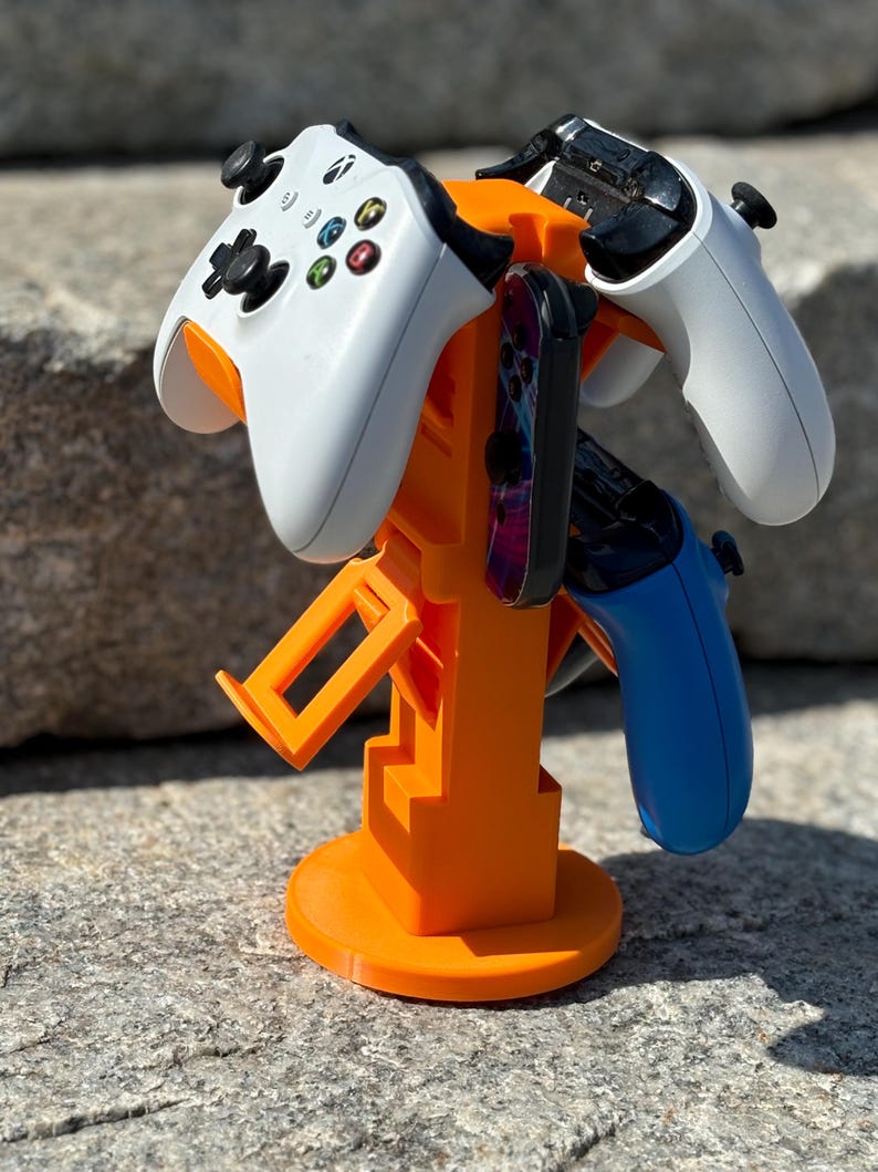 Puede incluir: Soporte de pl&aacute;stico naranja con tres mandos de videojuegos. El soporte tiene una base redonda y una estructura vertical con ranuras para sujetar los mandos. Los mandos son blancos, negros y azules.