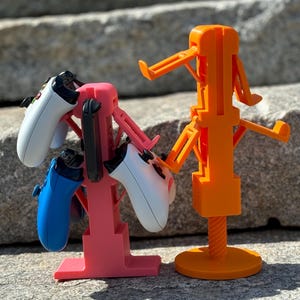 Puede incluir: Dos soportes impresos en 3D para mandos de videojuegos. El soporte de la izquierda es rosa y tiene dos mandos, uno azul y uno blanco. El soporte de la derecha es naranja y tiene un mando blanco.