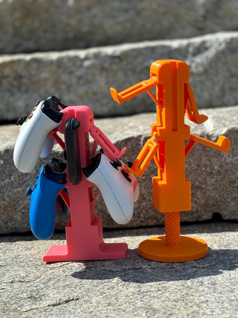 Puede incluir: Dos soportes impresos en 3D para mandos de videojuegos. El soporte de la izquierda es rosa y tiene dos mandos, uno azul y uno blanco. El soporte de la derecha es naranja y tiene un mando blanco.