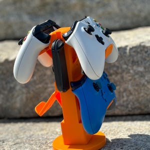 May include: Orange plastic stand with three slots for holding video game controllers. Two controllers are in the stand, one white and one blue.