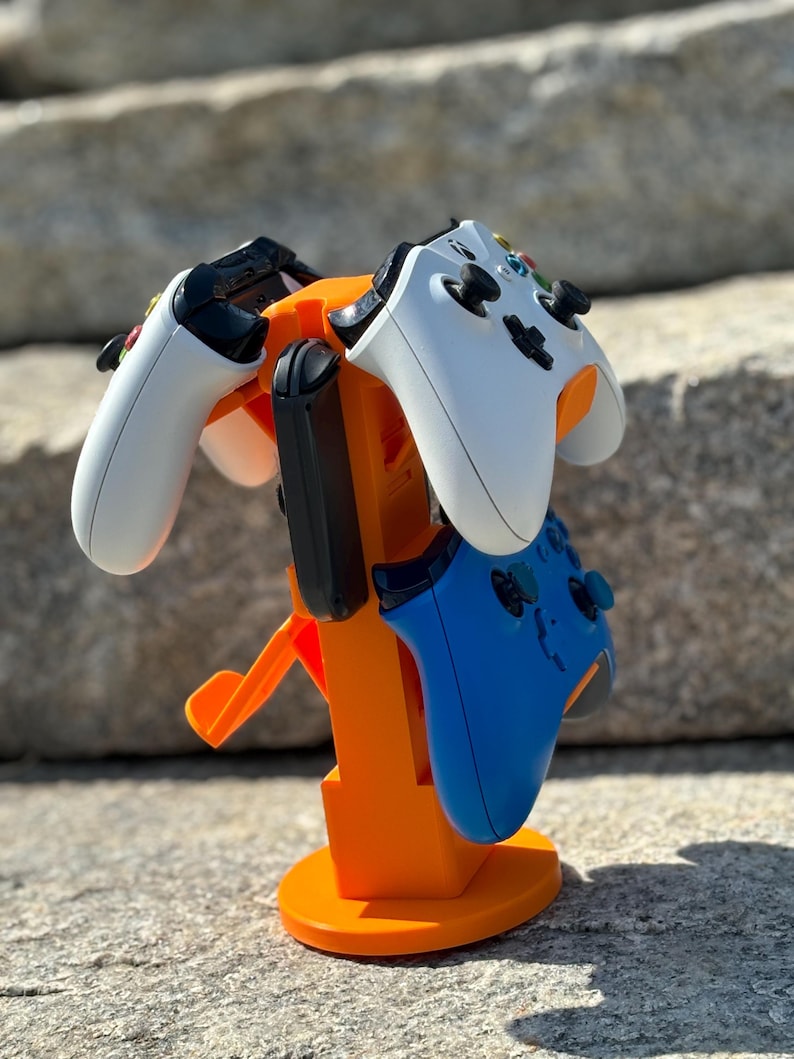 Puede incluir: Soporte de pl&aacute;stico naranja con tres ranuras para guardar mandos de videojuegos. Dos mandos est&aacute;n en el soporte, uno blanco y otro azul.