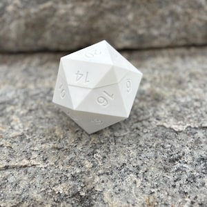 May include: A white, twenty-sided die, numbered from 1 to 20, rests on a gray, textured rock surface. The die is a solid, geometric shape with clearly defined edges and visible numbers. Ideal for tabletop games.