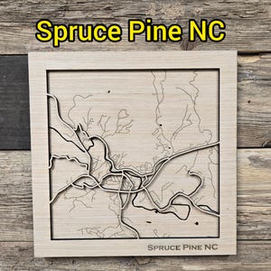 Könnte beinhalten: Eine Holzkarte von Spruce Pine, NC, mit einem detaillierten Laser-Cut-Design des Stadtlayouts. Die Karte ist in hellem Holz gerahmt, mit dem Text "Spruce Pine NC" oben und unten.