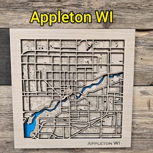 Puede incluir: Un mapa de Appleton, WI, cortado con láser en madera, con un río y un lago azules. La cuadrícula de calles de la ciudad está intrincadamente detallada. El texto "Appleton WI" está en la parte superior e inferior del mapa. El mapa está sobre un fondo de madera rústica.