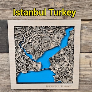 May include: A wooden map of Istanbul, Turkey, with a blue water background. The map is laser-cut with intricate details of the city's streets and buildings. The text "Istanbul Turkey" is displayed at the top and bottom of the map.