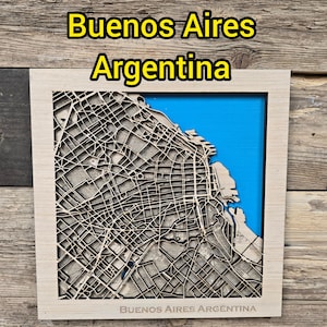 May include: A wooden map of Buenos Aires, Argentina, with a detailed laser-cut design. The map is set against a blue background, framed in light wood, and includes the text "Buenos Aires Argentina".