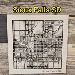 May include: A light-colored wooden map of Sioux Falls, SD, with intricate laser-cut details of streets and waterways. The city name is displayed in yellow at the top and bottom of the frame. The map is set against a rustic wood background.