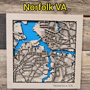 May include: A wooden map of Norfolk, VA, with a light wood frame. The map features intricate laser-cut streets and waterways, with a blue background representing the water. The text "Norfolk VA" is at the top and bottom of the frame.