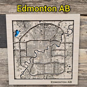 Op de afbeelding: Een houten kaart van Edmonton, Alberta, met de tekst "Edmonton AB" bovenaan en onderaan. De kaart is ingewikkeld uitgesneden en toont straten, waterwegen en een blauw meer. Het frame is van lichtgekleurd hout.