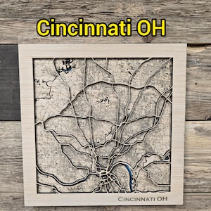 Op de afbeelding: Een houten kaart van Cincinnati, Ohio, met een licht houten frame. De kaart toont gedetailleerde straten en waterwegen. De naam van de stad staat in het geel bovenaan en onderaan het frame.