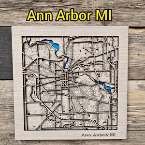 Könnte beinhalten: Eine Holzkarte von Ann Arbor, MI, mit einem detaillierten Laser-Cut-Design. Die Straßen und Wasserwege der Stadt sind aufwendig geschnitzt, mit blauen Akzenten für die Wasserelemente. Der Text "Ann Arbor MI" ist oben und unten angezeigt.