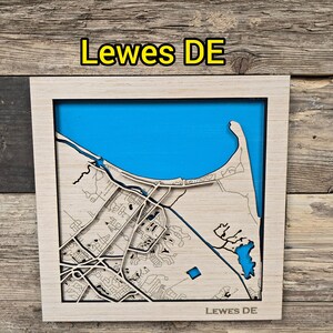 May include: A wooden map of Lewes, Delaware, with a light wood frame. The map features a detailed laser-cut design of the city's layout, with a blue background representing the water. The text "Lewes DE" is at the top and bottom.