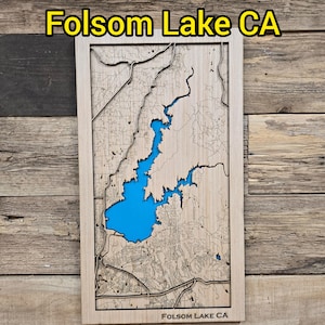 May include: A wooden map of Folsom Lake, CA, with a blue lake area. The map is framed in a light wood color and has the text "Folsom Lake CA" at the top and bottom. The map is mounted on a wooden background.