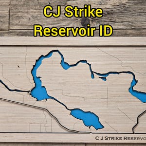 May include: Wooden map of CJ Strike Reservoir ID, with a light wood grain background and a blue reservoir. The map is framed in a light wood frame. The text "CJ Strike Reservoir ID" is displayed at the top and bottom of the map.