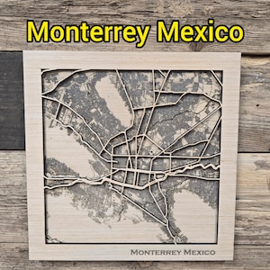 May include: A wooden map of Monterrey, Mexico, with a detailed laser-cut design. The city's layout is visible in gray, set within a light-colored wooden frame. The text "Monterrey Mexico" is displayed at the top and bottom of the frame.